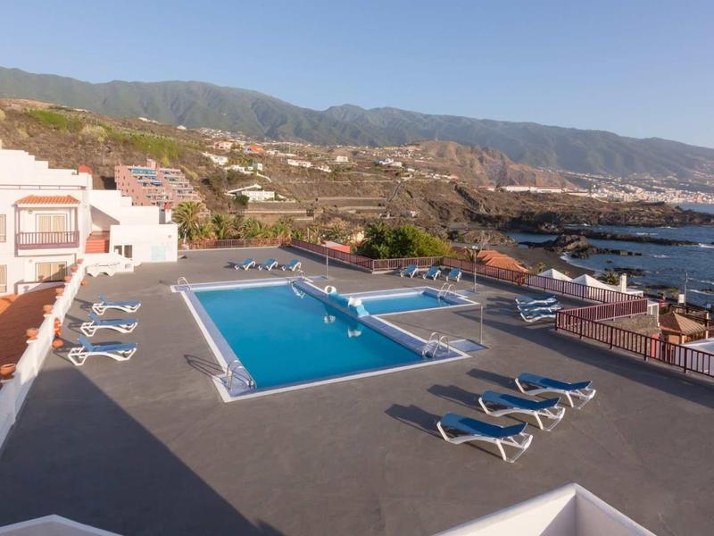 Hotel pool with sun loungers and view of the sea and mountains under clear sky.