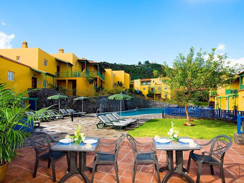 Acogedora zona exterior con mesas, sillas y una piscina frente a edificios amarillos bajo un cielo azul.