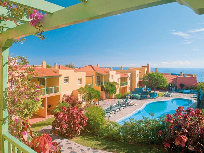 Vista desde el balcón a la piscina y casas en fila con vista al mar bajo cielo despejado.
