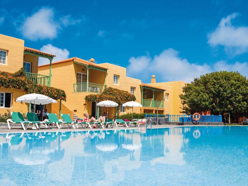 Hotel con edificio amarillo, sombrillas y tumbonas junto a una gran piscina clara bajo cielo azul.