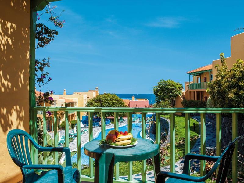 Balcón con mesa y sillas verdes, vista a palmeras y mar bajo cielo azul.