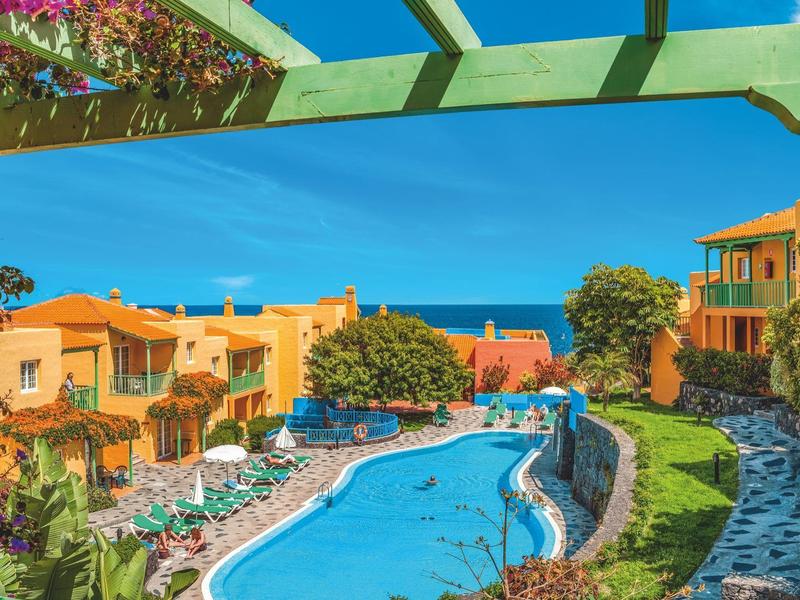 Piscina al aire libre rodeada de tumbonas y edificios coloridos bajo un cielo despejado.