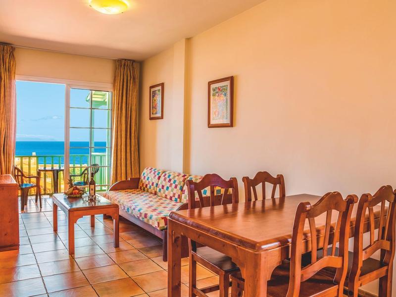 Sala luminosa con muebles de madera, sofá y vista al mar por puertas de balcón abiertas.