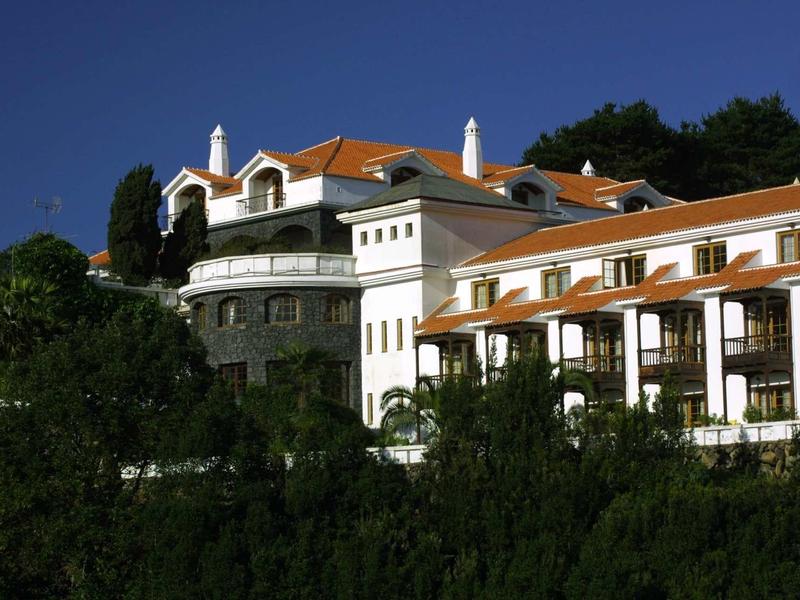 Gran edificio de hotel con paredes blancas y techos de tejas rojas frente a un cielo azul y árboles verdes.