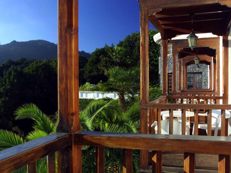 Balcón de madera con vistas a un paisaje montañoso y palmeras bajo un cielo azul claro.