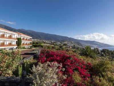 Hotel con balconi accanto a un giardino con fiori rossi davanti a montagne e mare.