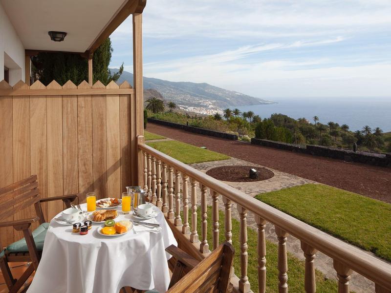 Balkon hotelowy z nakrytym stołem, widokiem na morze i zadbanym ogrodem.