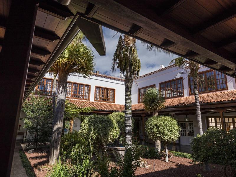 Dziedziniec hotelowy z palmami, oknami z okiennicami i czerwonym dachem z dachówek.