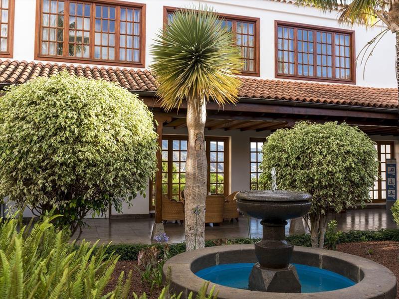 Fontanna otoczona drzewami i krzewami przed budynkiem w stylu kolonialnym.