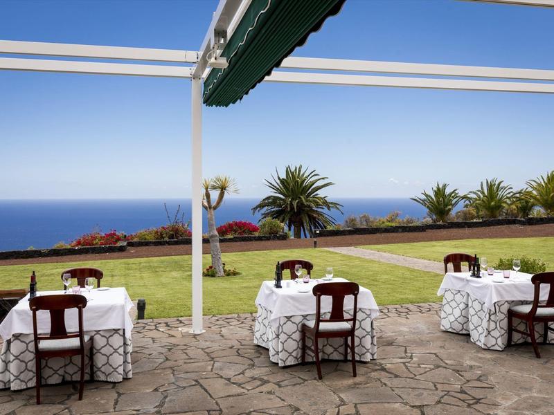 Taras z stołami, krzesłami, widokiem na ogród i morze w tle.