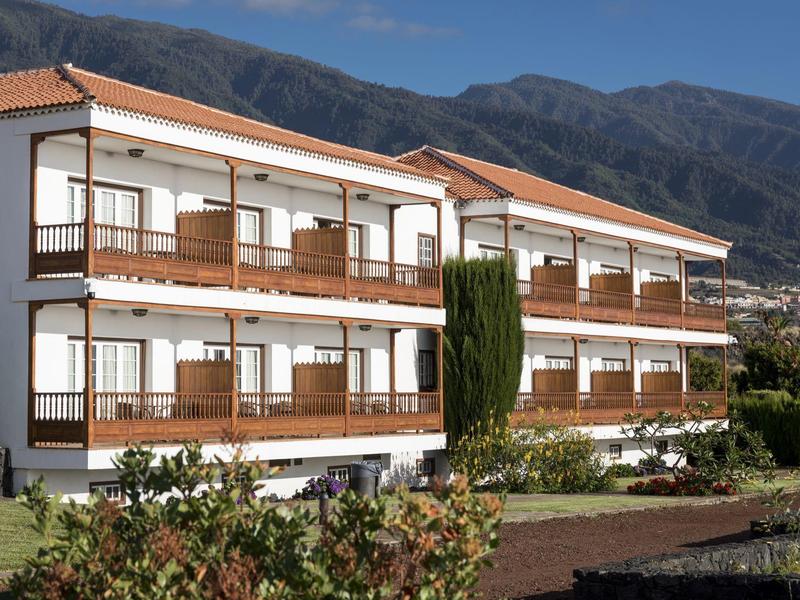 Biały hotel z drewnianymi balkonami na tle gór.