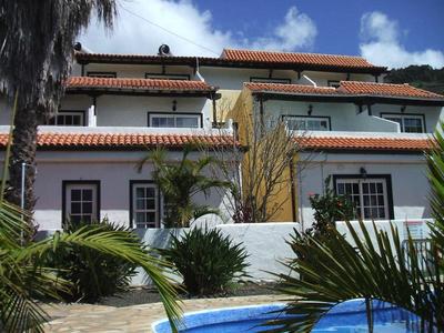 Zwei weiße Häuser mit roten Ziegeldächern, grünen Fensterläden und Palmen im Vordergrund.