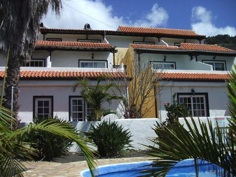 Twee witte huizen met rode daken en planten ervoor onder een heldere lucht.