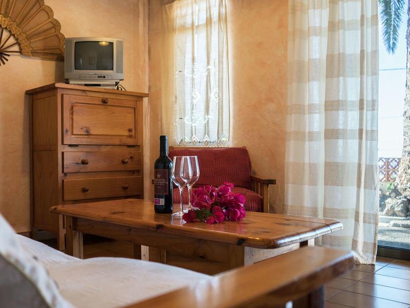 Pohodlná část hotelového pokoje s dřevěným nábytkem, lahví vína, sklenicemi a květinami na stole.