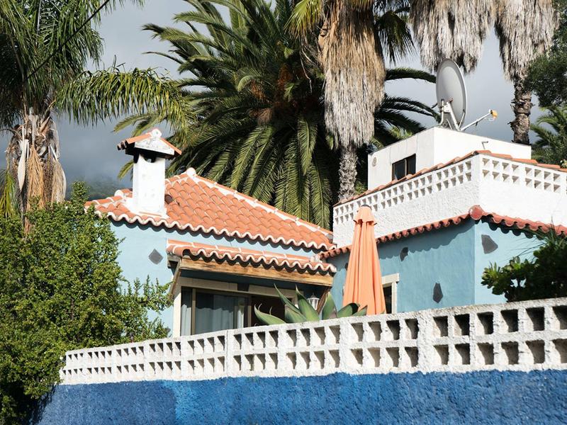 Modrý dům s červenými taškami a palmami v pozadí pod oblačnou oblohou.