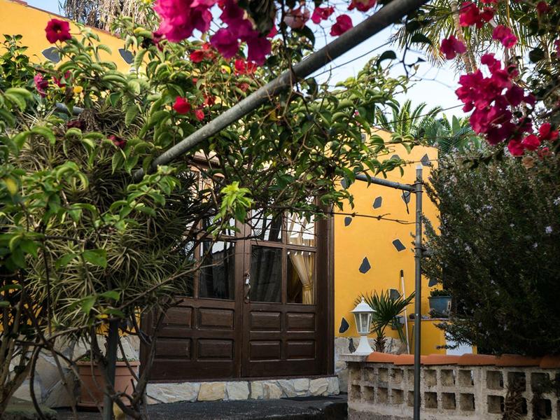 Kvetoucí rostliny rámují posezení se židlemi před žlutou budovou.