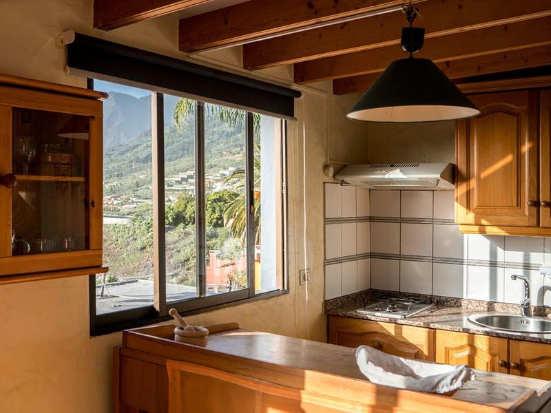 Malá kuchyň s dřevěným nábytkem, velké okno s výhledem na hory a viditelné stropní trámy.