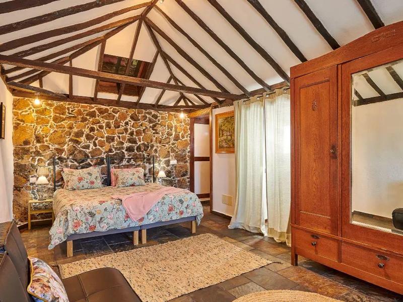 Pohodlná ložnice s dřevěnými trámy, kamenným pozadím a starožitnou dřevěnou skříní.