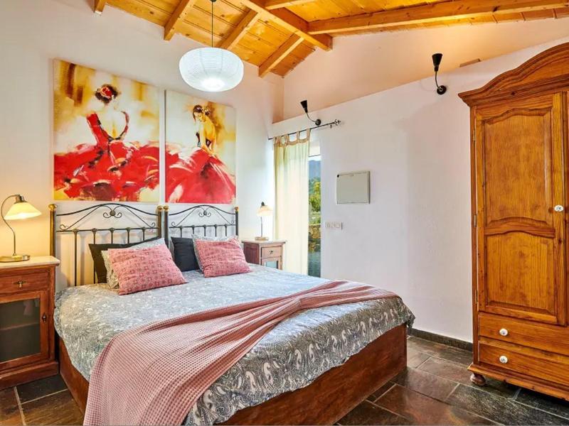 Pohodlná ložnice s dřevěnou postelí, růžovými doplňky a starožitnou dřevěnou skříní pod dřevěným stropem.