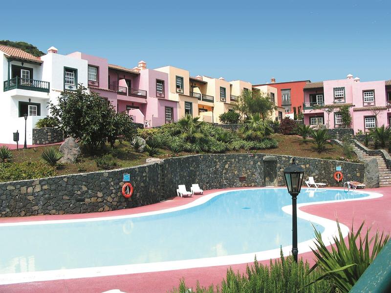 Mehrfarbige Reihenhäuser oberhalb eines klaren, geschwungenen Schwimmbeckens bei sonnigem Himmel.