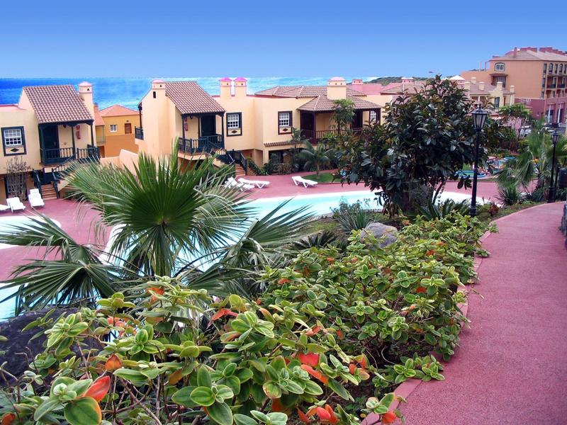 Bunte Gartenzone mit Palmen und Sträuchern neben einem roten Gehweg und Häusern im Hintergrund.