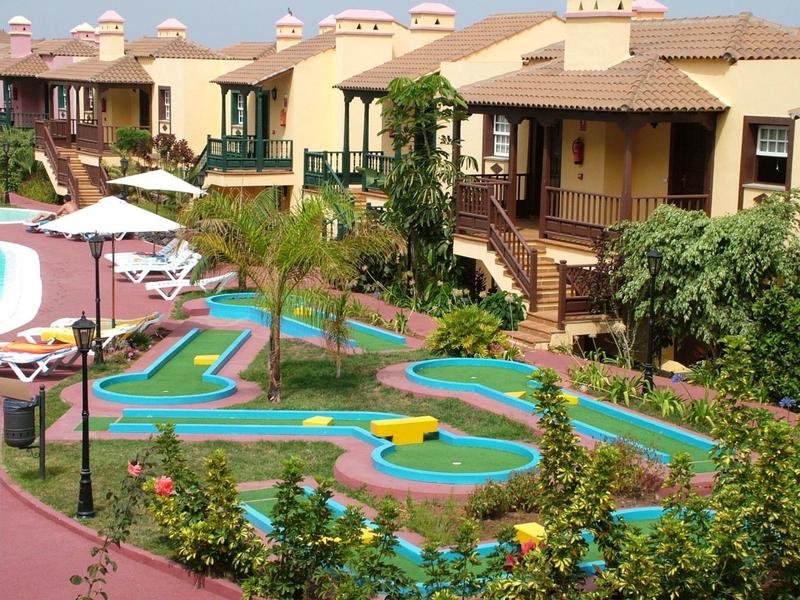 Ein buntes Minigolfspiel vor braunen Ferienhäusern mit Palmen und grünen Pflanzen.