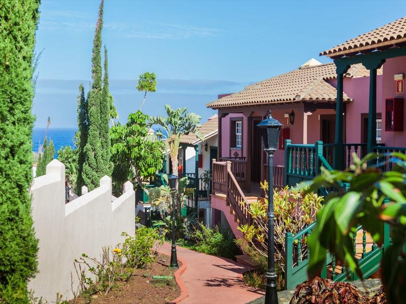 Mediterranes Haus mit roten Wänden, grünem Geländer, Gartenweg und blauem Meer im Hintergrund.