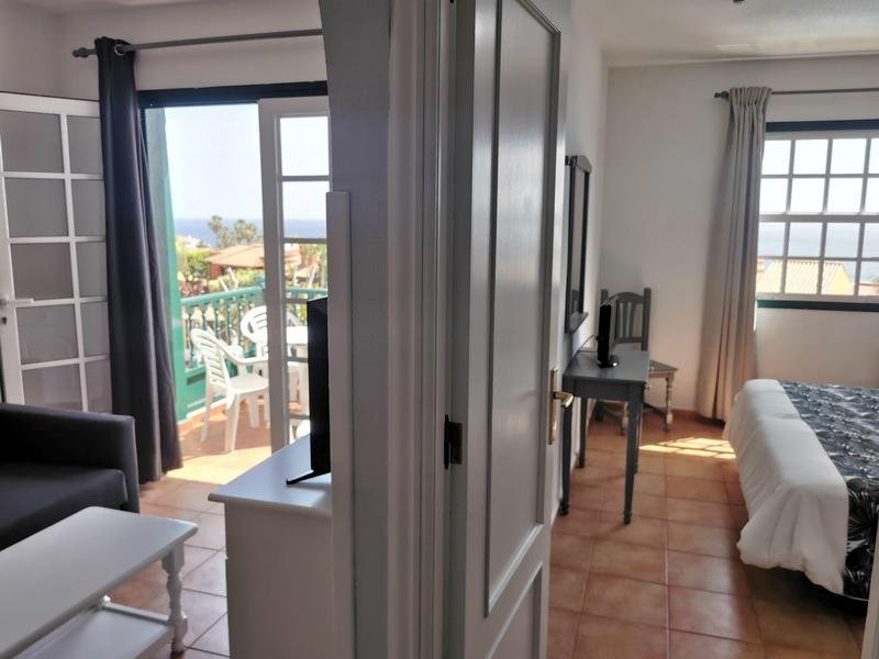 Helles Zimmer mit Fliesenboden, Balkon, Couch, Schreibtisch und großem Fenster mit Vorhängen.