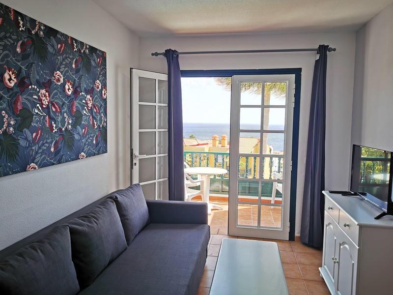 Wohnzimmer mit grauem Sofa, Balkon mit Meerblick, blauem Vorhang und buntem Wandbild.
