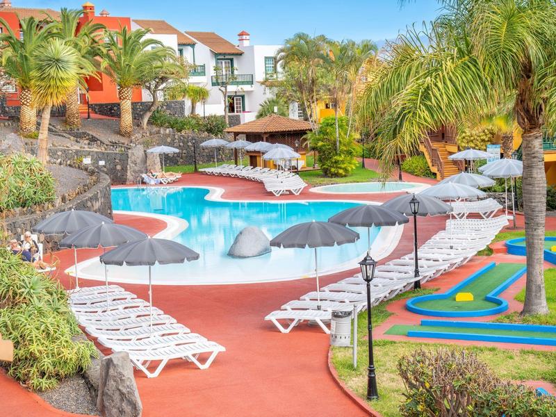 Schwimmbadbereich mit Liegestühlen, Sonnenschirmen, Palmen und buntem Minigolfplatz im Freien.