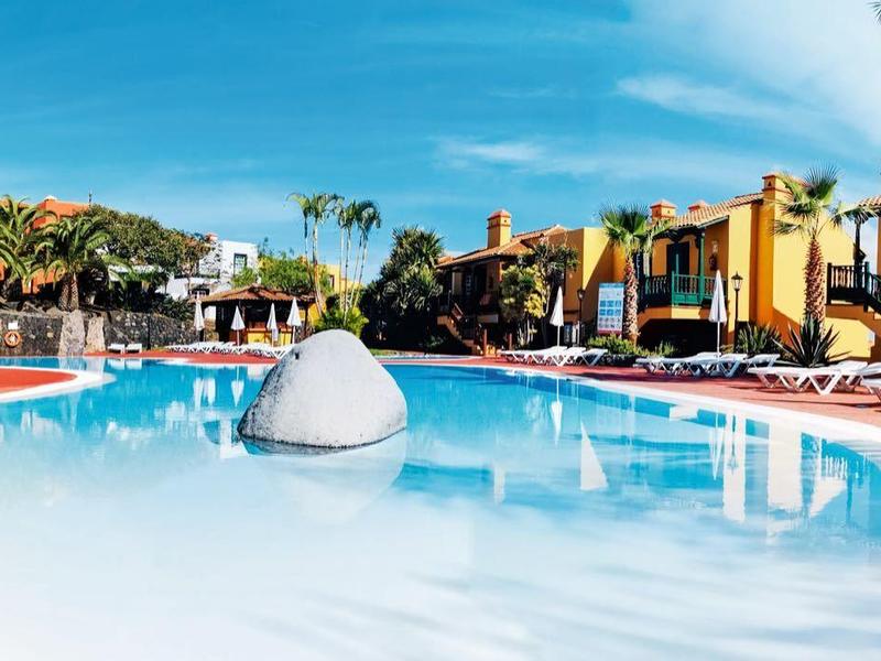 Großer Pool mit klarem Wasser, umgeben von Palmen und gelben Bungalows unter blauem Himmel.