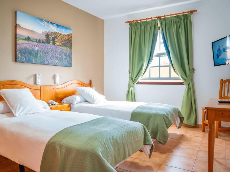 Hotelzimmer mit zwei Einzelbetten, grünem Vorhang und einfachem Holzmobiliar.