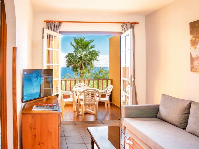 Helles Wohnzimmer mit Couch, TV und Blick auf Balkon mit Palmen und Meer.
