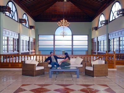 Amplia sala de estar de hotel con vista al mar, sofá y huéspedes conversando.