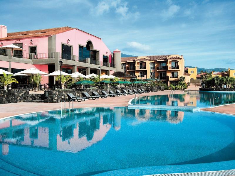 Gran piscina exterior con tumbonas frente a hoteles de varios pisos bajo un cielo azul.