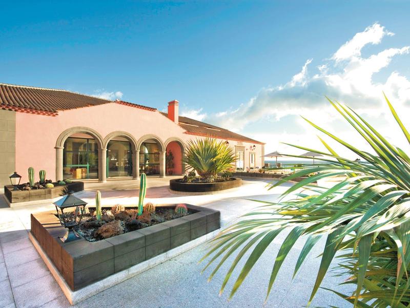 Amplia terraza con muebles lounge y plantas verdes frente a un edificio de hotel en día soleado.