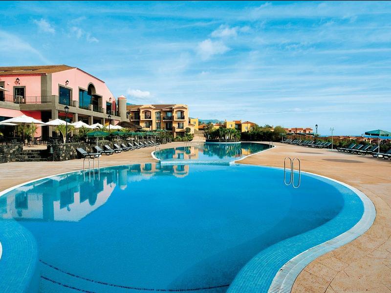 Gran piscina frente a un hotel con tumbonas y cielo azul.