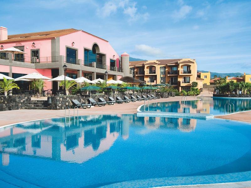 Gran piscina con tumbonas y sombrillas frente a un hotel bajo un cielo azul.