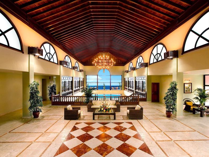 Amplio vestíbulo de hotel con techo de madera, suelo en cuadros y vista al mar al fondo.