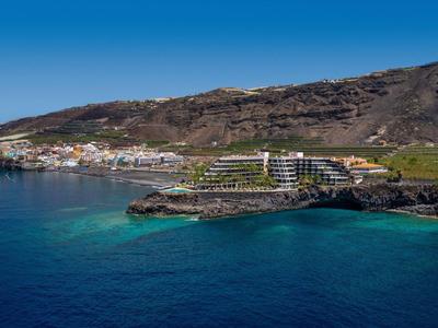 Hotel by the sea on a rocky coast with clear blue water and mountains in the background.