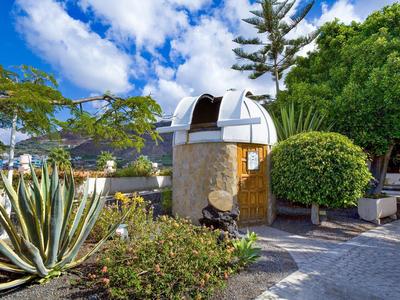 Small stone building with silver metal roof in a tropical garden under blue sky.