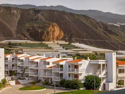 Modern white apartments with red roofs set against mountainous landscape under cloudy sky.