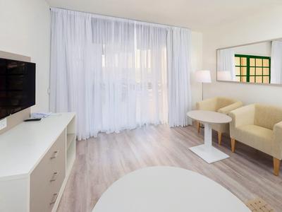 Bright hotel room with TV, two armchairs, table, and large windows with curtains.