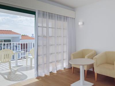 Bright room with two armchairs, small table, and balcony with plastic chair overlooking rooftops.