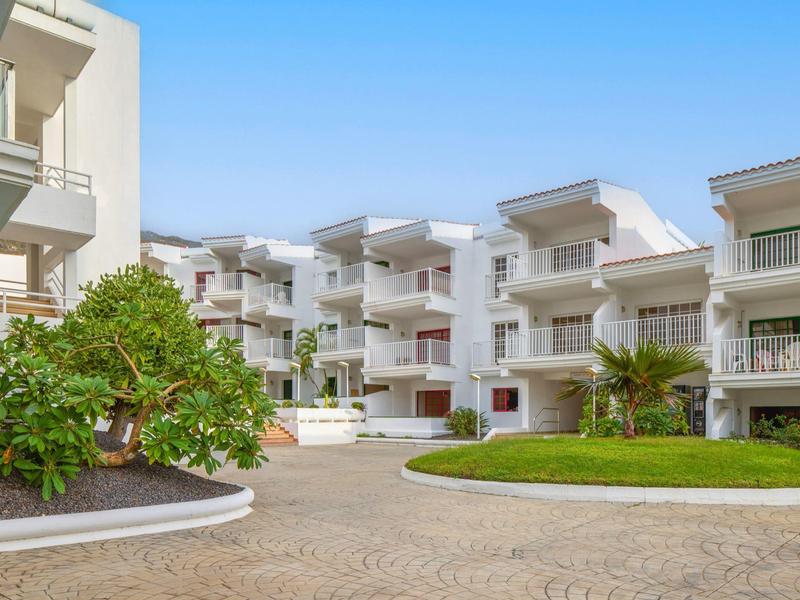Modern white residential buildings with balconies and well-kept garden under clear sky.