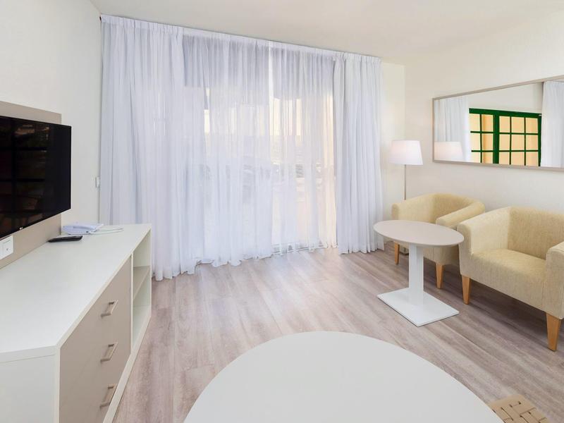 Bright hotel room with TV, two armchairs, table, and large windows with curtains.