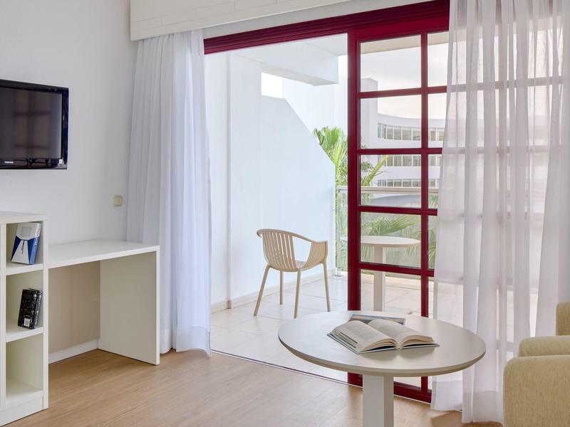 Bright hotel room with round table, two chairs, and balcony door with red railing.