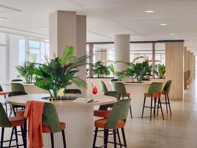 Espace commun lumineux et moderne avec tabourets de bar, chaises vertes et plantes.