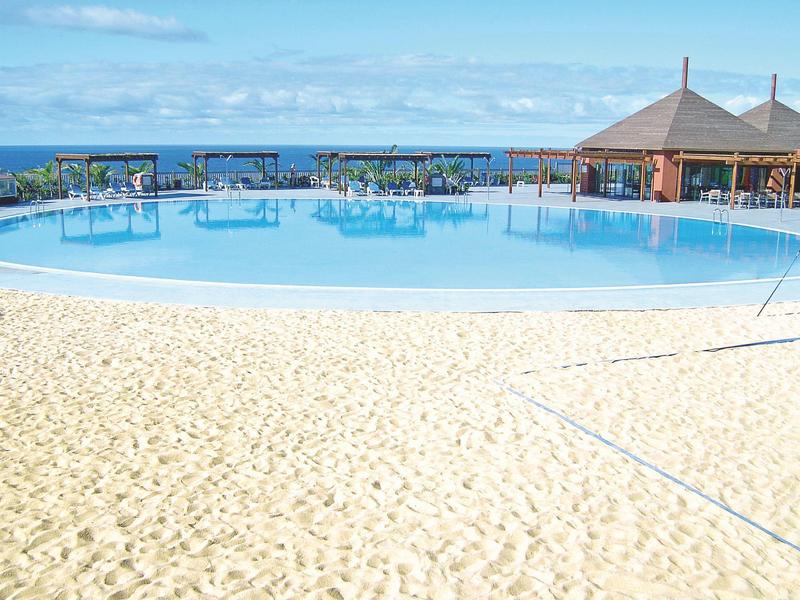 Großer blauer Pool mit runder Form, umgeben von Sand und einer Bar mit Sonnenschirmen am Meer.