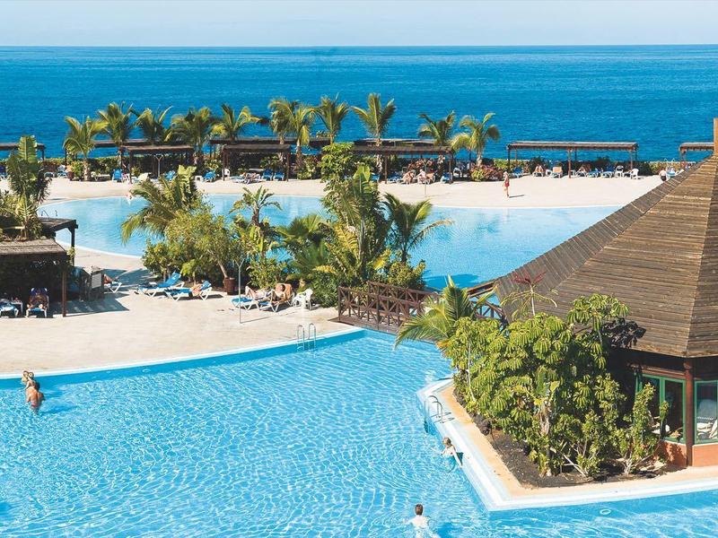 Blick auf großes blaues Schwimmbecken mit Palmen, Sandstrand und Meer im Hintergrund bei Sonnenschein.